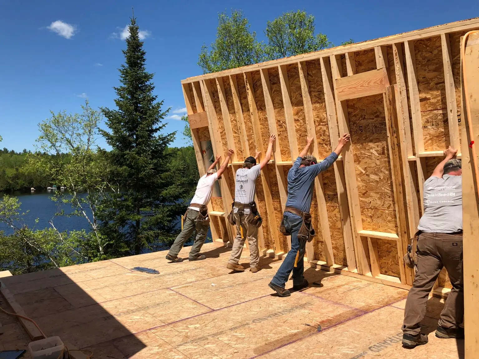 Four construction workers raising a wooden wall frame on a building site, with a river and trees visible in the background under a clear blue sky. Lakeside Construction Management is a professional construction project oversight and construction management firm specializing in construction oversight and cost control