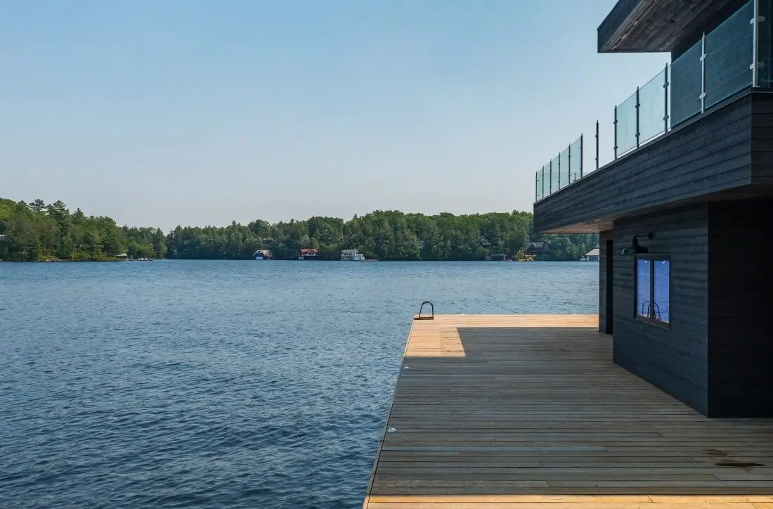A wooden dock extending over a calm lake, with a modern building featuring large windows in the foreground. Lush green trees line the opposite shore, under a clear blue sky, exemplifying a serene setting suitable for construction project oversight or cottage construction management in Ontario. Lakeside Construction Management is a professional construction project oversight and construction management firm specializing in construction oversight and cost control